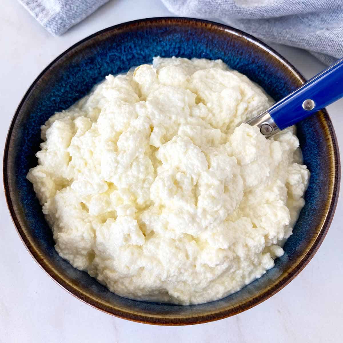 A blue bowl filled with homemade ricotta cheese and a blue spreading knife.