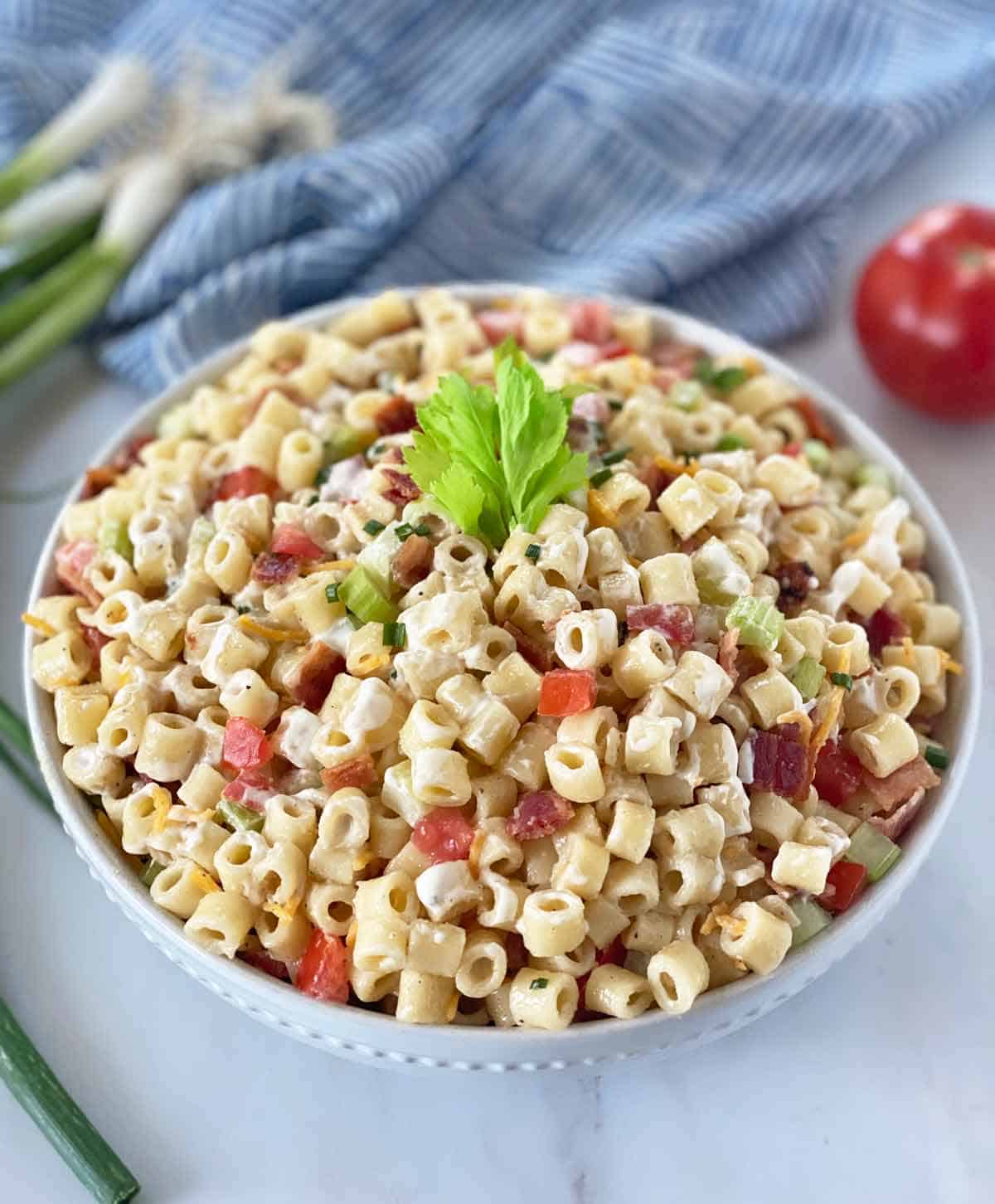 Macaroni salad with bacon in a white serving bowl with scallions and a tomato in the background.