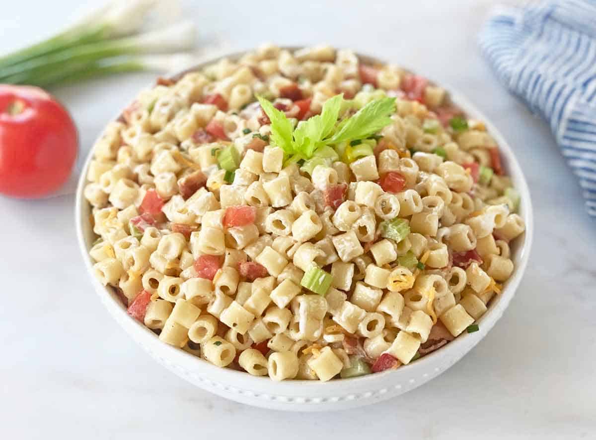 A white shallow serving bowl filled with bacon macaroni salad with vegetables in the background.