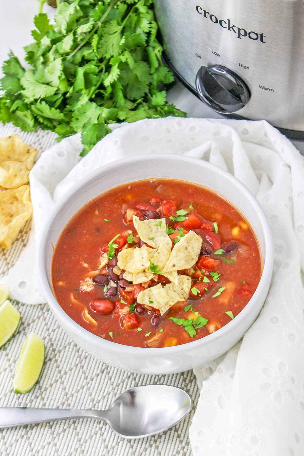 A white bowl with Southwest Chicken Chili and cilantro and a crock pot in the background.