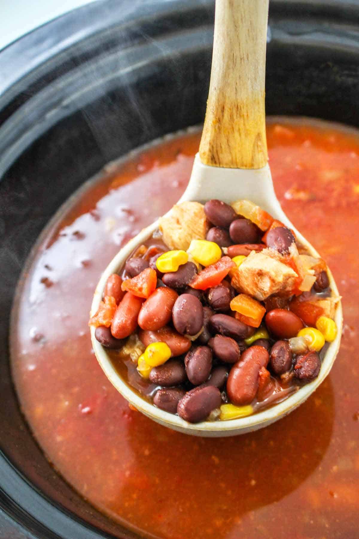 A ladle filled with cooked chicken chili with beans, corn, chicken and chipotle peppers.