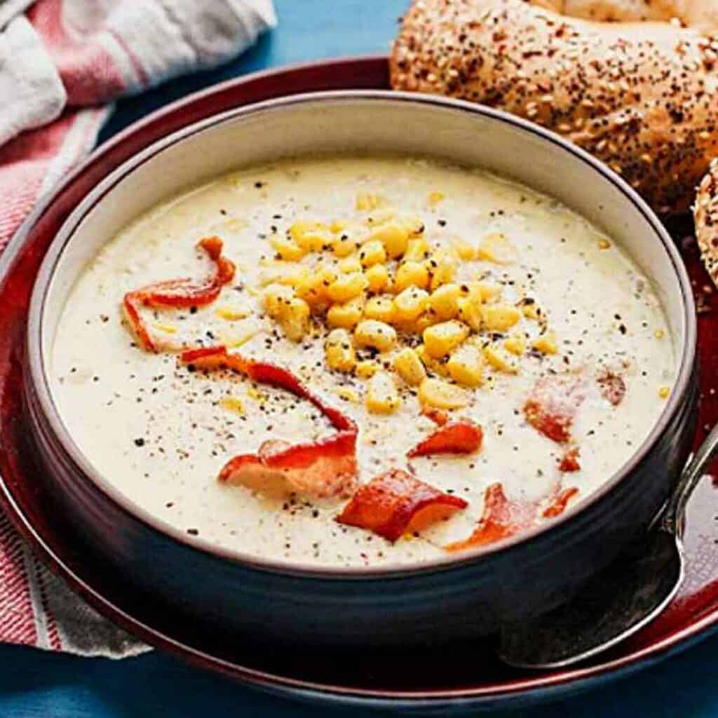 A ceramic bowl with old fashioned corn chowder with bacon, and a bagel on the plate.