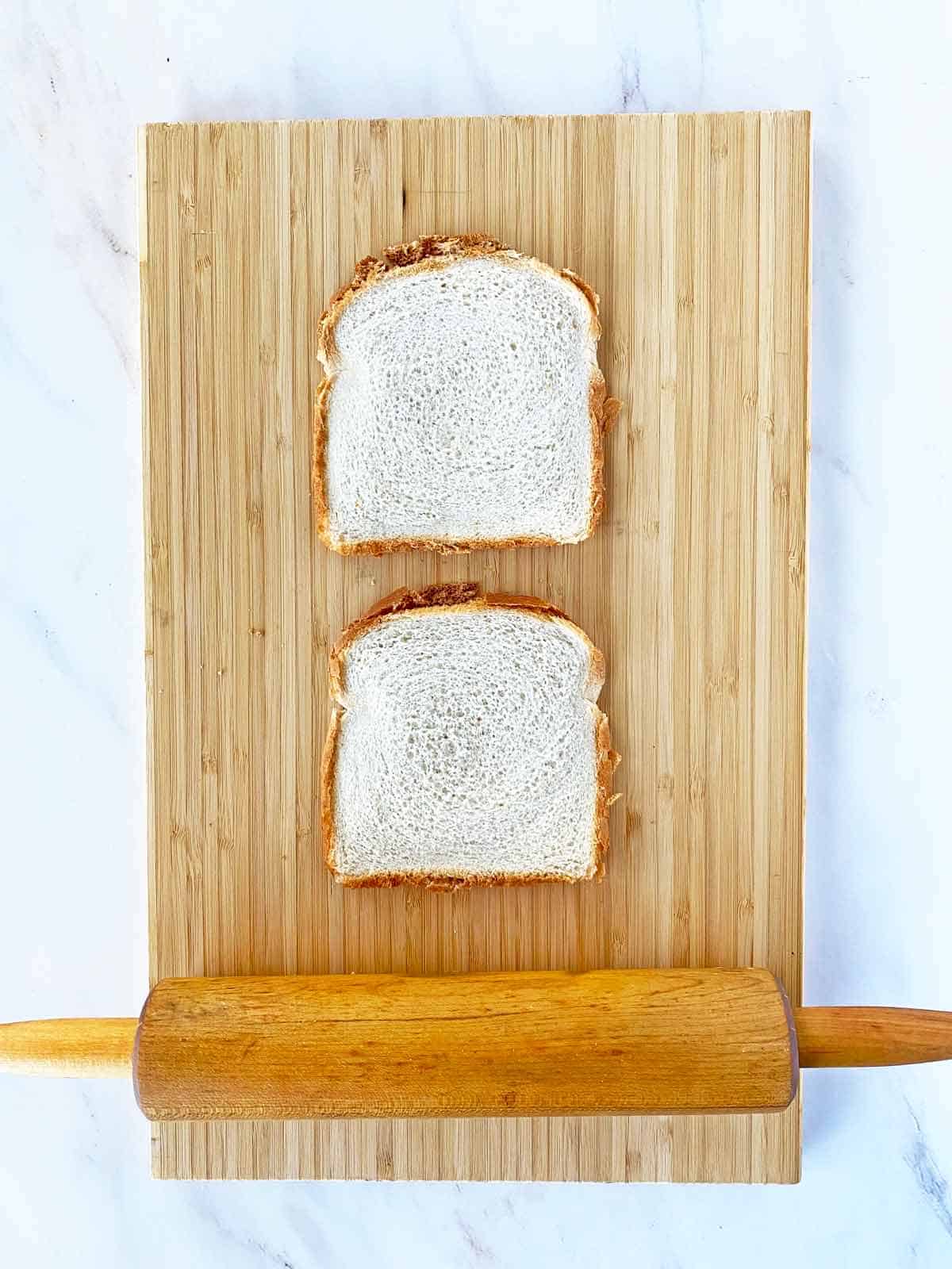 Two pieces of bread being flattened by a rolling pin on a cutting board.