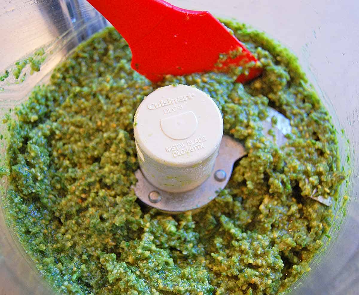 Scraping down the pesto in a food processor with a red spatula.